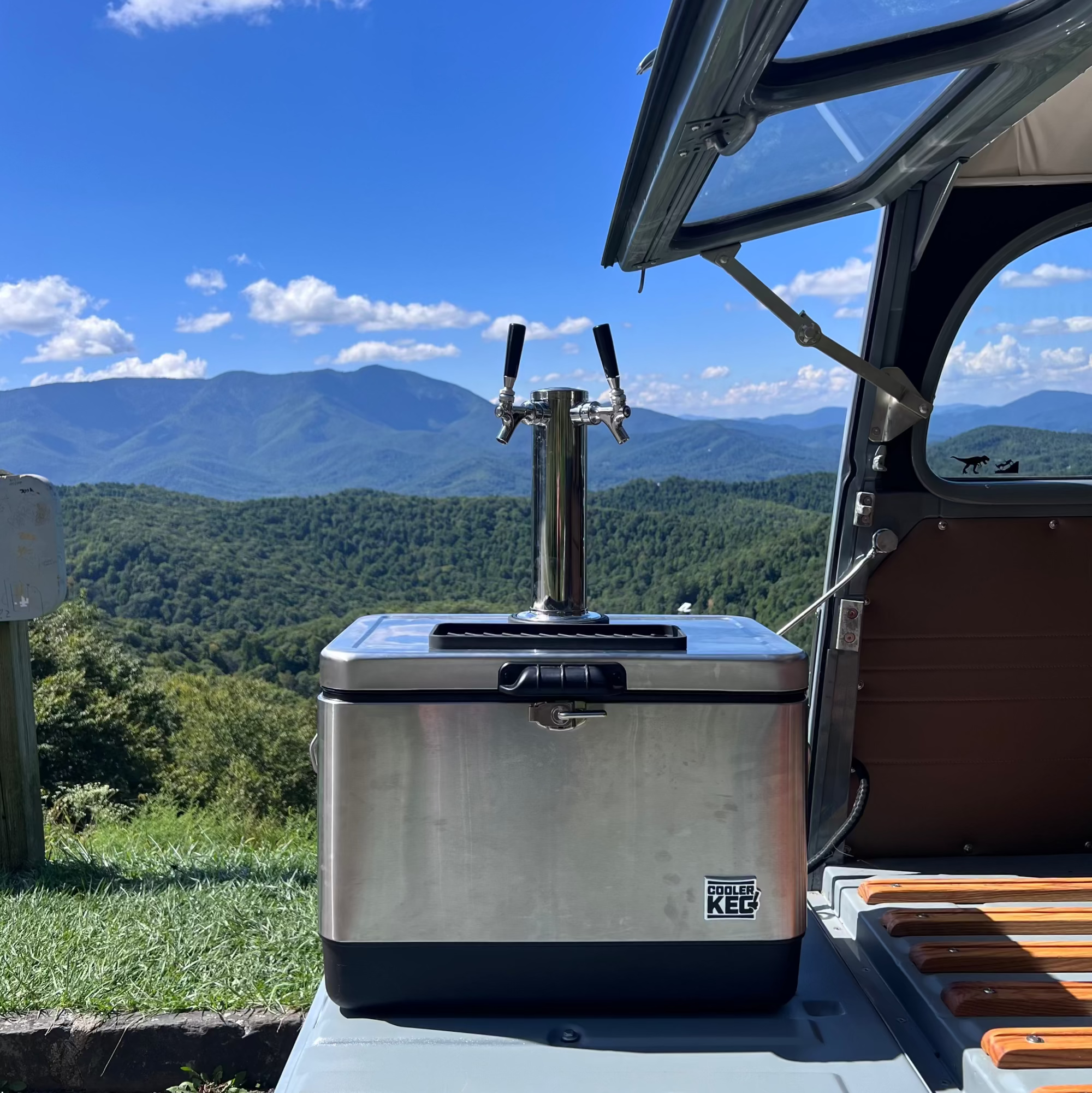 Stainless steel cooler with a tap on a vehicle's open roof, with mountains in the background.