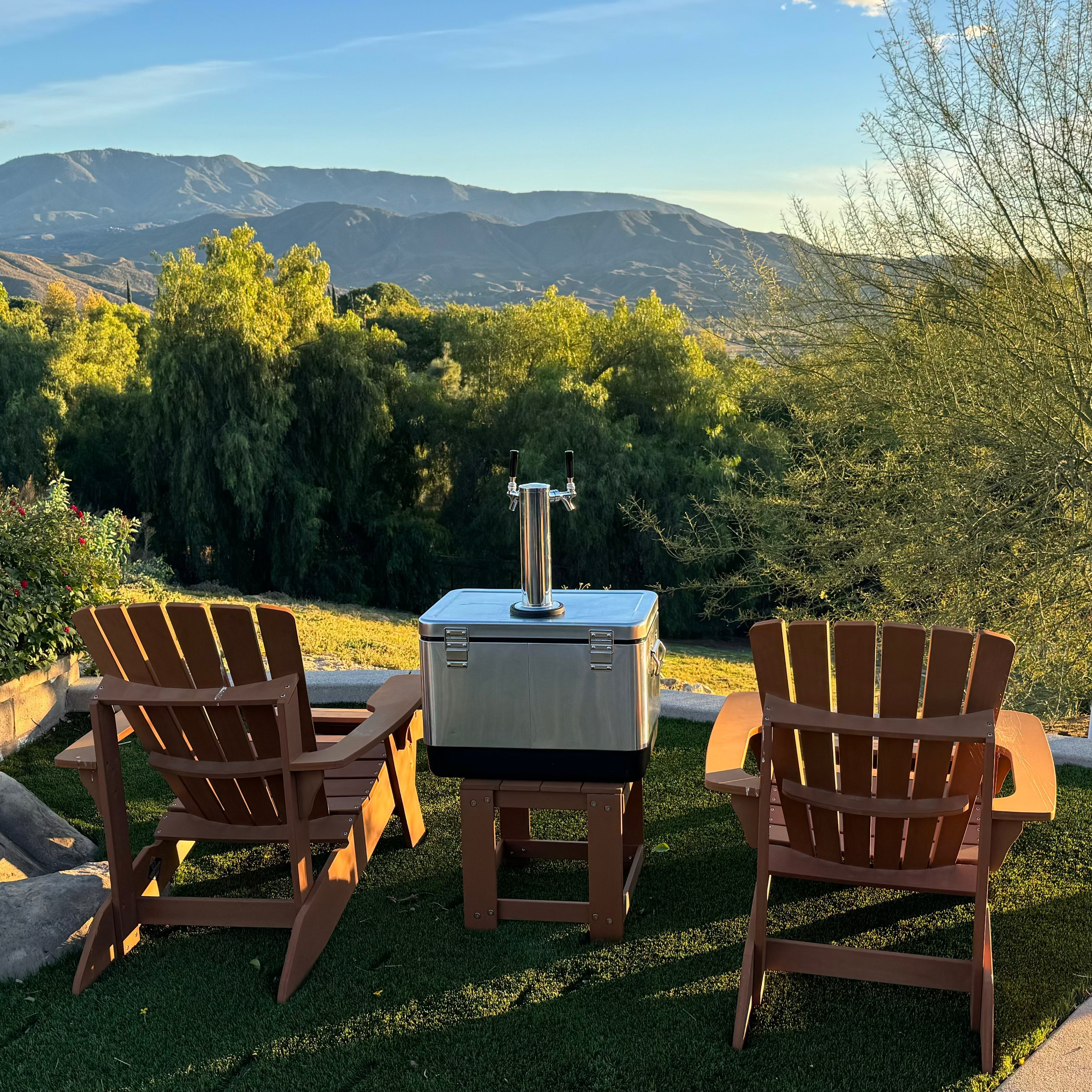 Two wooden chairs and a small table with a grill on a grassy area with mountains in the background.