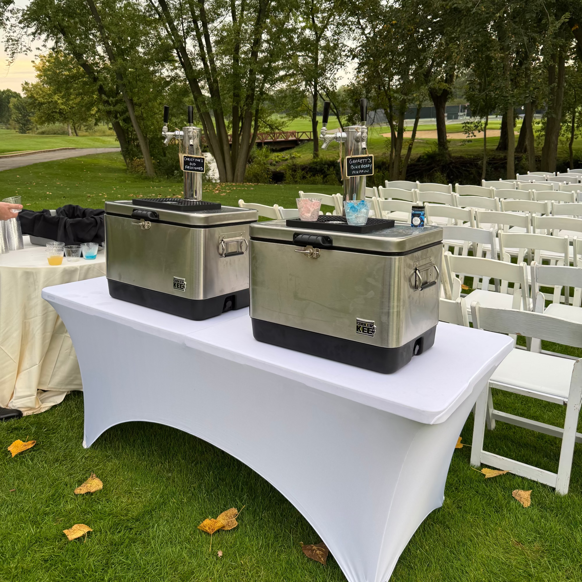 Two large metal coolers on a white table outdoors with trees and chairs in the background.