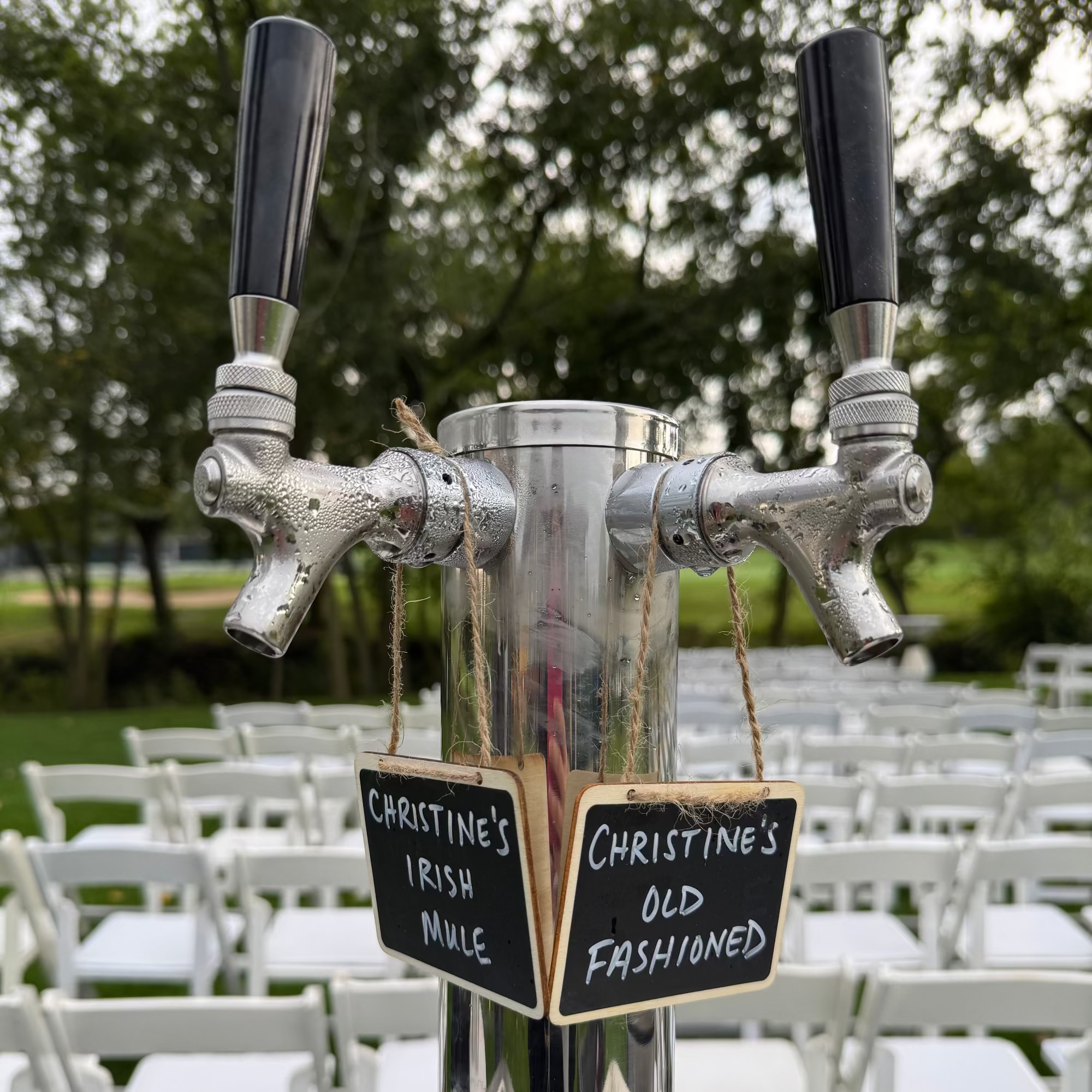 Metallic drink dispenser with two taps labeled 'Christine's Irish Mule' and 'Christine's Old Fashioned' in an outdoor setting.