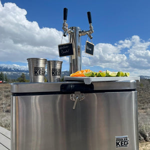 Stainless steel beer keg with two taps and a tray of fruit under a blue sky with clouds.